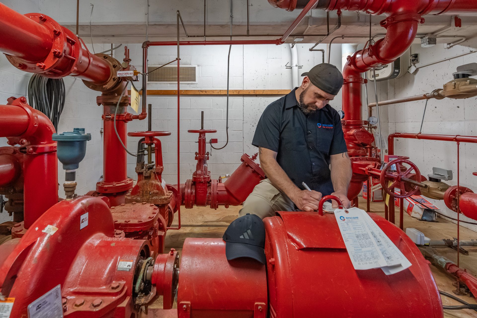 Encore technician performing a fire pump inspection, documenting results on-site in a mechanical room with red fire protection system piping and equipment.