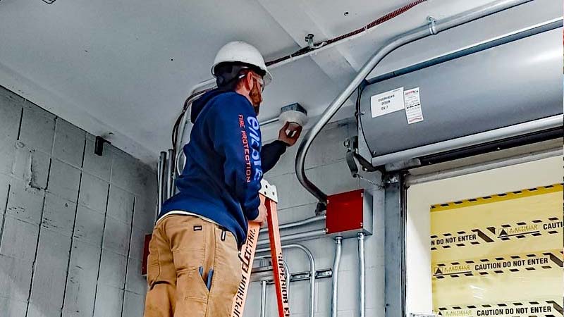 Encore technician in a hard hat and harness inspecting a fire alarm system on a ladder in a facility
